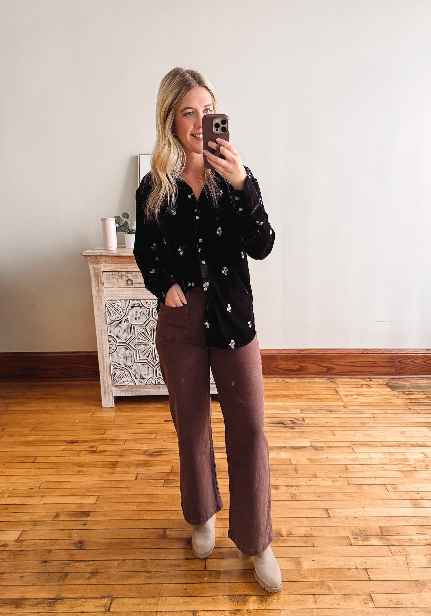 Woman taking a mirror selfie wearing a black patterned shirt and brown pants in a room with wooden flooring.