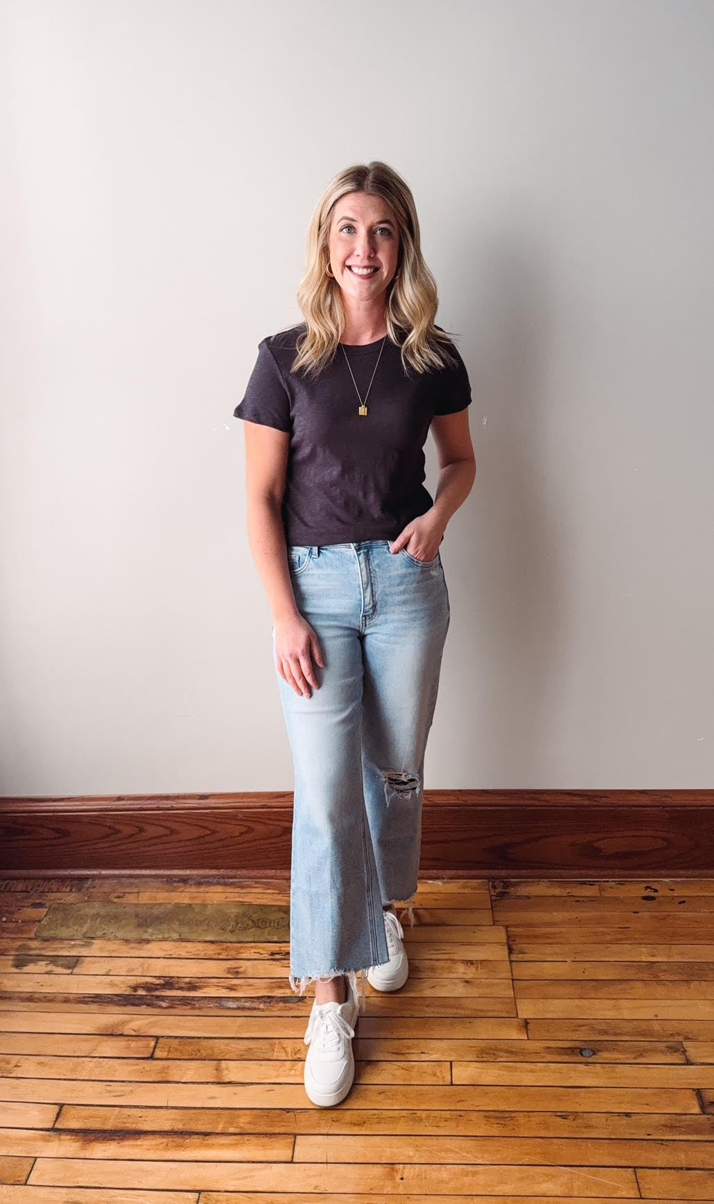 Woman wearing a black t-shirt, light blue jeans, and white sneakers standing on a wooden floor.