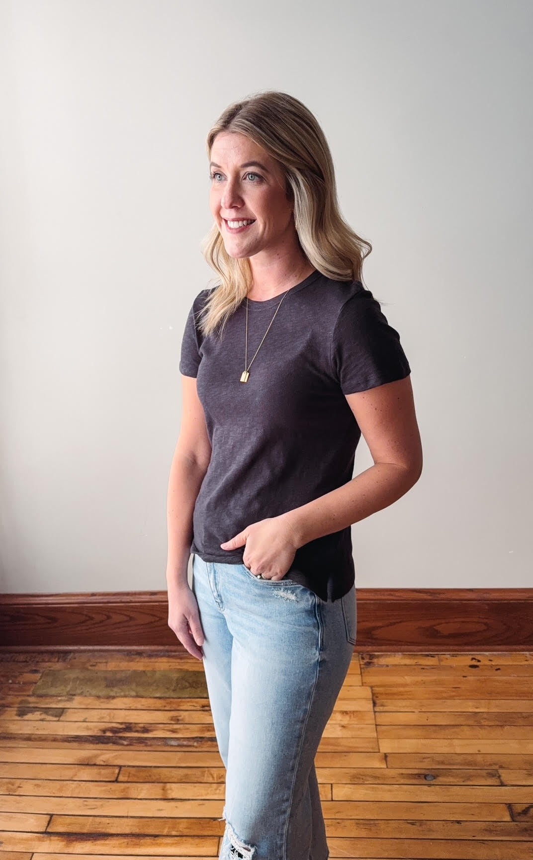 Woman wearing a dark gray t-shirt and light blue jeans standing on a wooden floor.