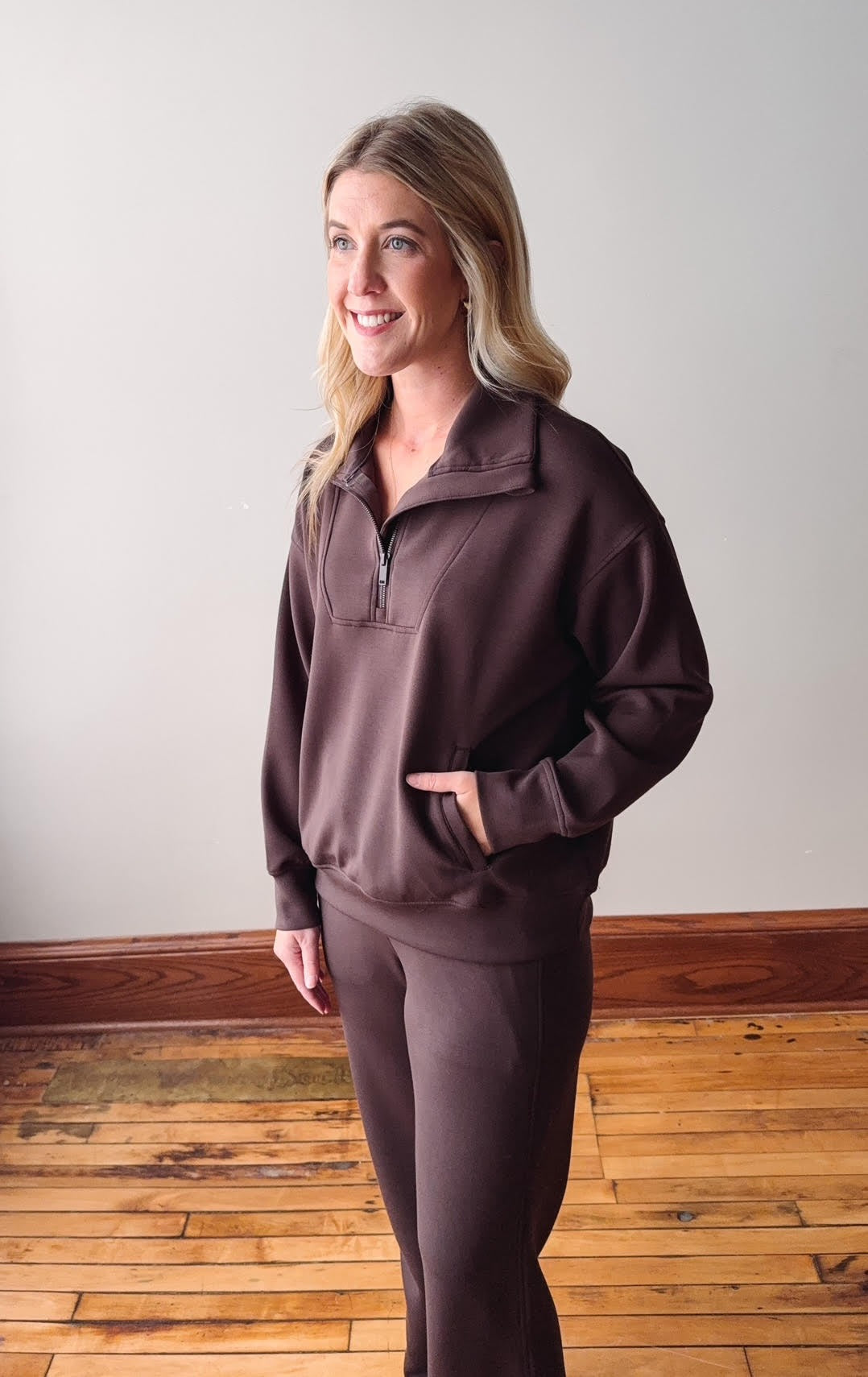 Woman wearing a brown tracksuit standing on a wooden floor with a white wall background