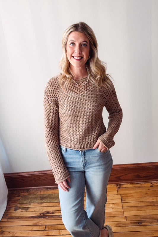 Woman wearing a beige knitted sweater and light blue jeans standing against a white wall.