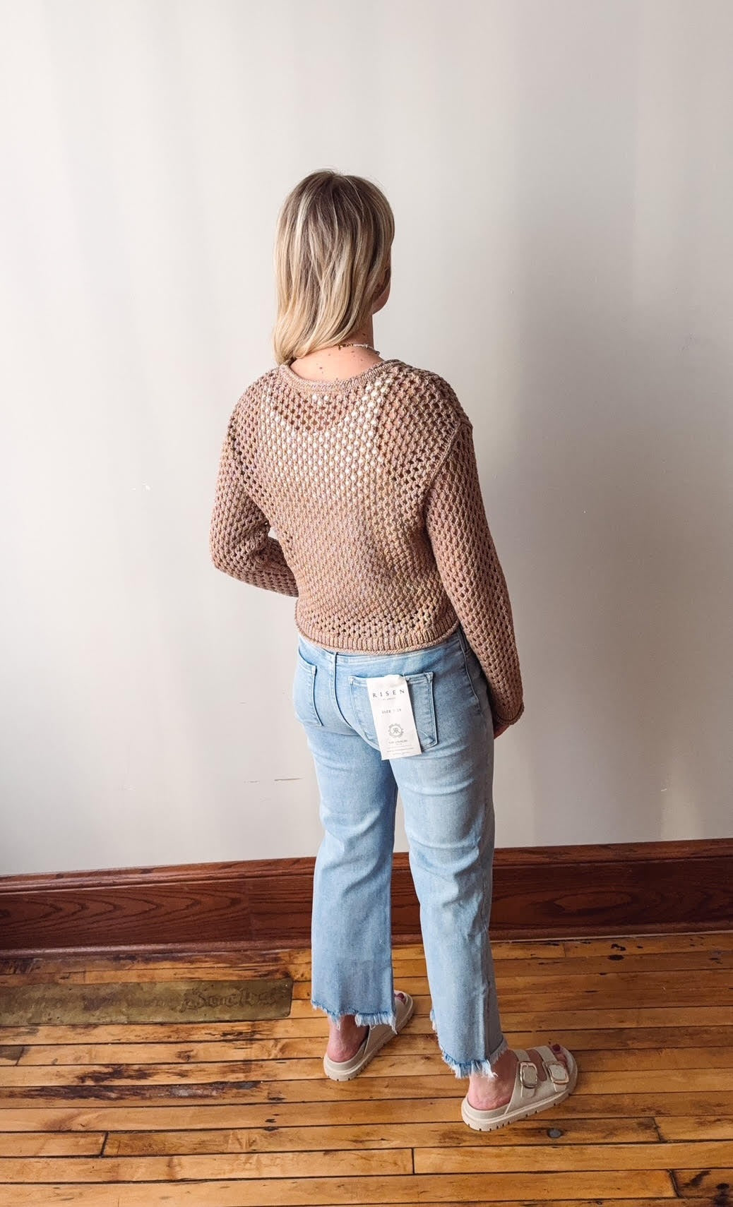 Person wearing a sheer brown top and blue jeans standing against a white wall.