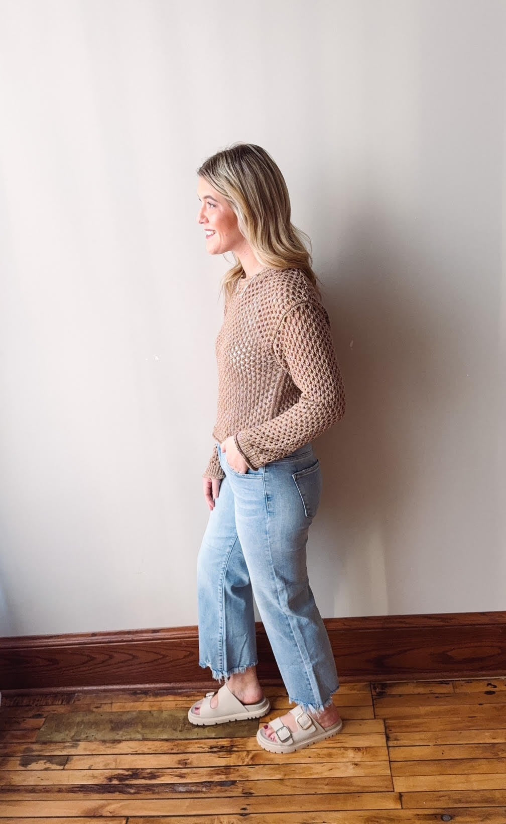 Woman wearing a brown textured sweater and light blue jeans standing against a white wall.