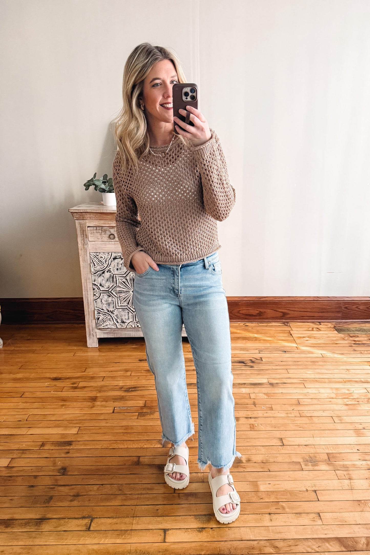 Woman taking a mirror selfie wearing a brown sweater and light blue jeans in a room with wooden flooring.