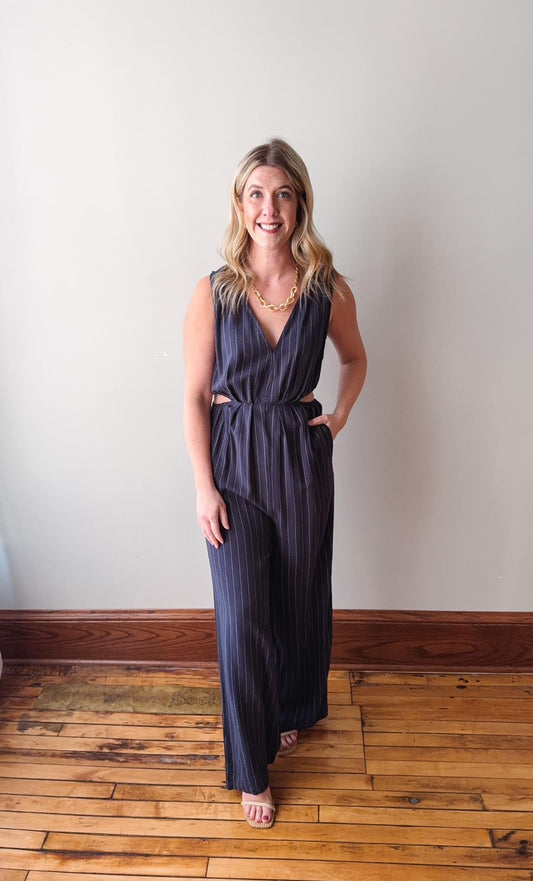 Woman wearing a dark blue jumpsuit standing on a wooden floor against a white wall.