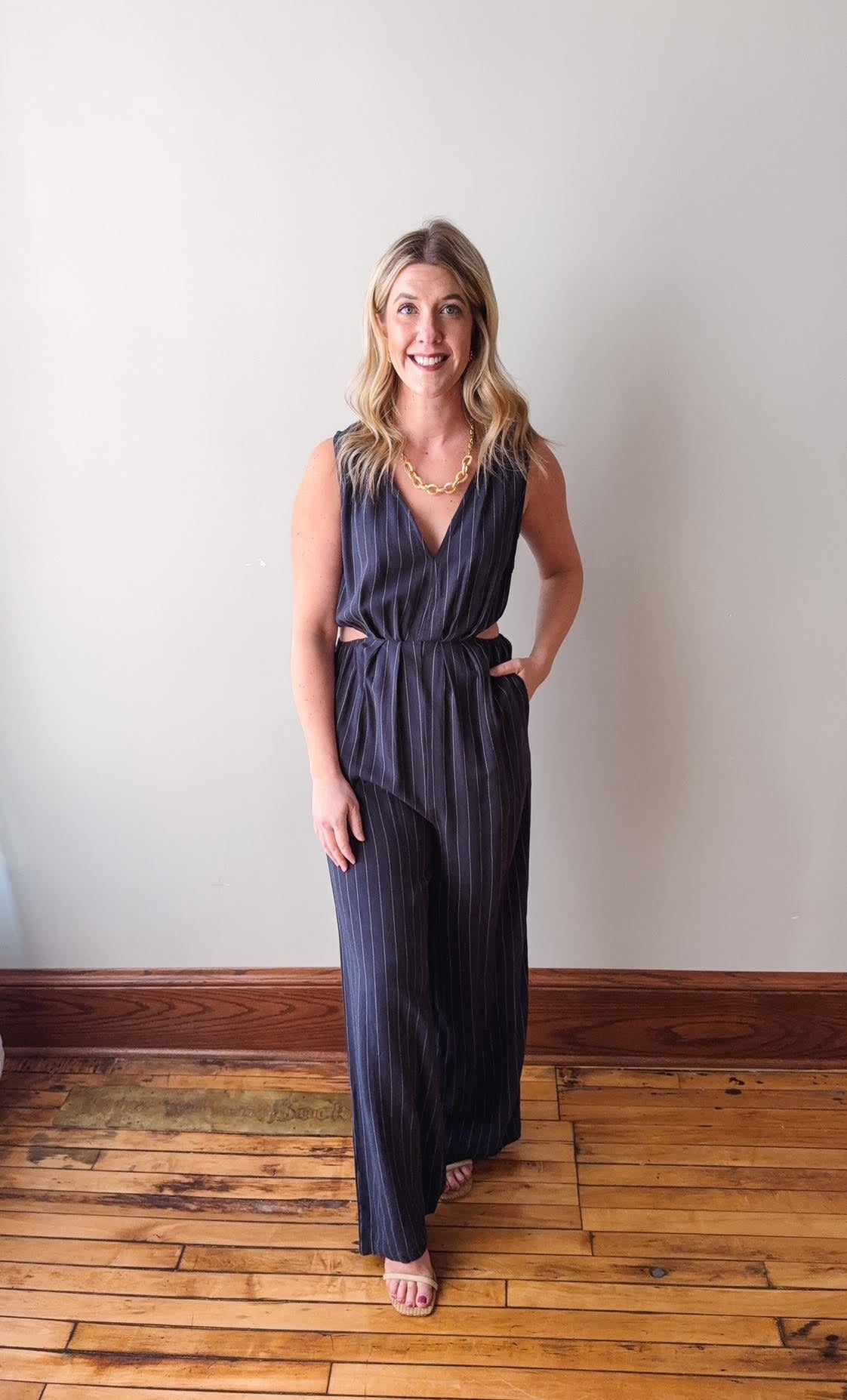 Woman wearing a dark blue jumpsuit standing on a wooden floor against a white wall.