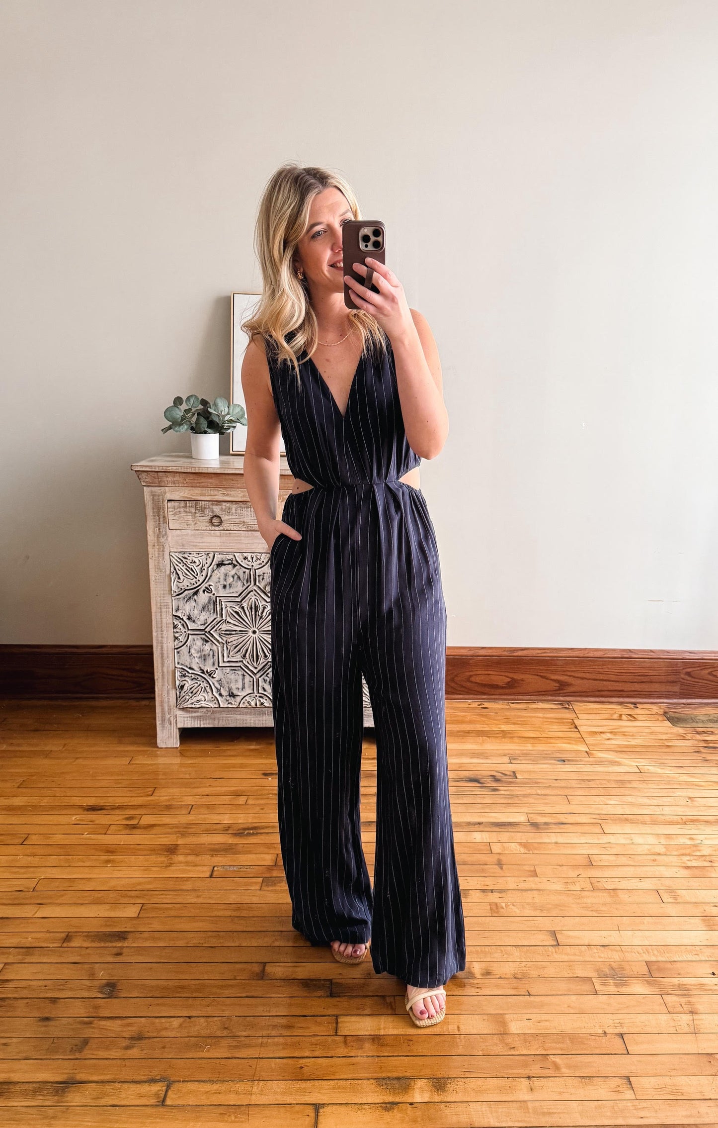 Woman taking a mirror selfie wearing a navy jumpsuit in a room with wooden flooring and a decorative cabinet.