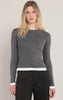 A woman modeling a dark gray long-sleeve crewneck sweater with contrasting white ribbed trim at the collar, hem, and cuffs, paired with black trousers against a light beige studio background.