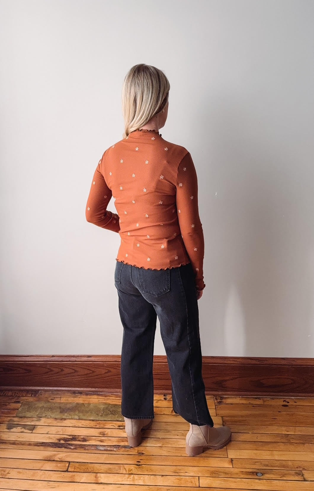 Person wearing an orange patterned shirt and dark pants standing on a wooden floor against a white wall.