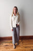 Woman wearing a white blouse and gray pants standing against a plain wall.