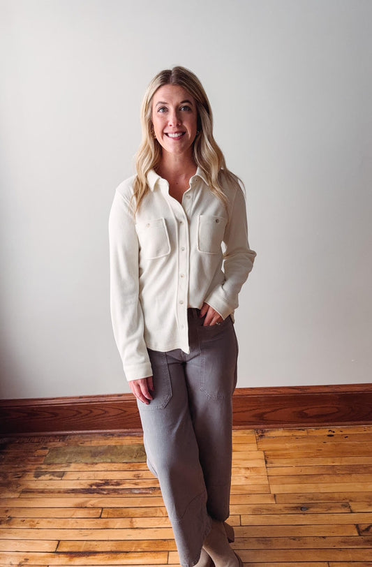 Woman wearing a white shirt and gray pants standing in a room with wooden flooring and a white wall.
