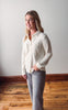 Woman wearing a white blouse and gray pants standing on a wooden floor with a white wall background
