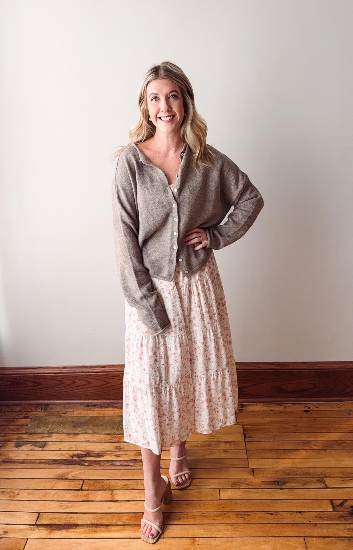 Woman wearing a gray cardigan and floral dress standing on a wooden floor with a white wall background