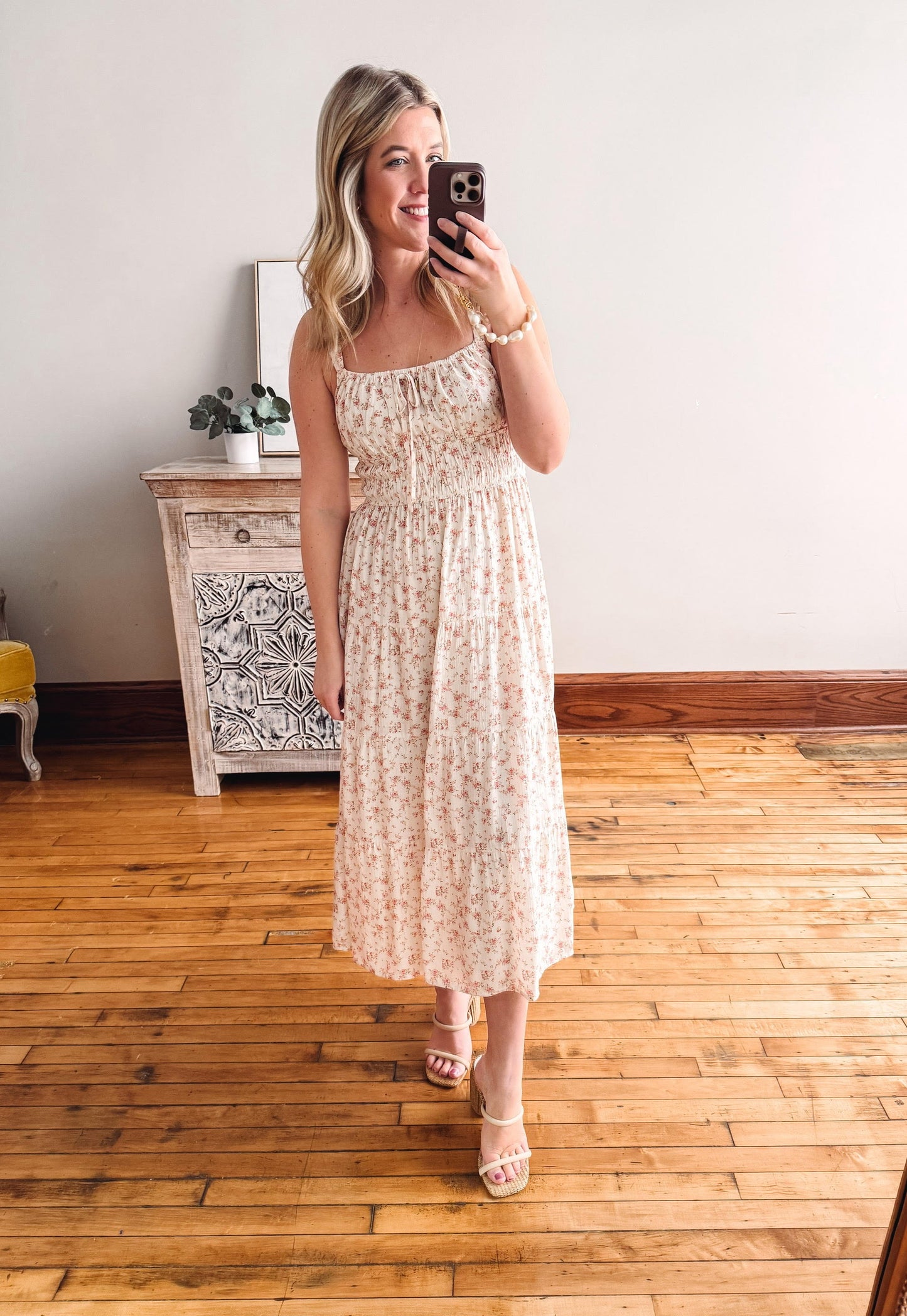 Woman taking a mirror selfie wearing a floral dress in a room with wooden flooring.