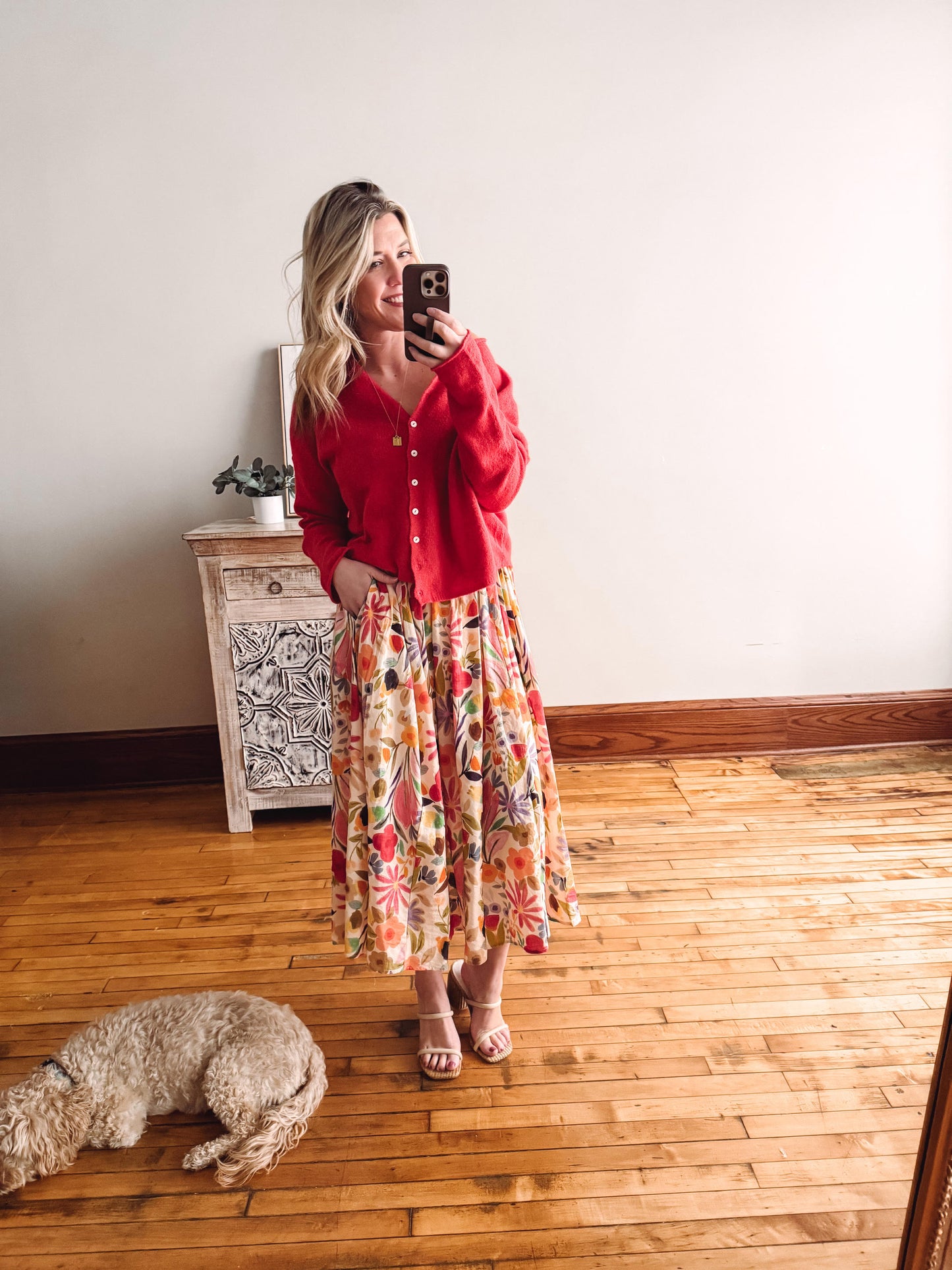 Woman taking a mirror selfie wearing a red shirt and floral skirt on a wooden floor.