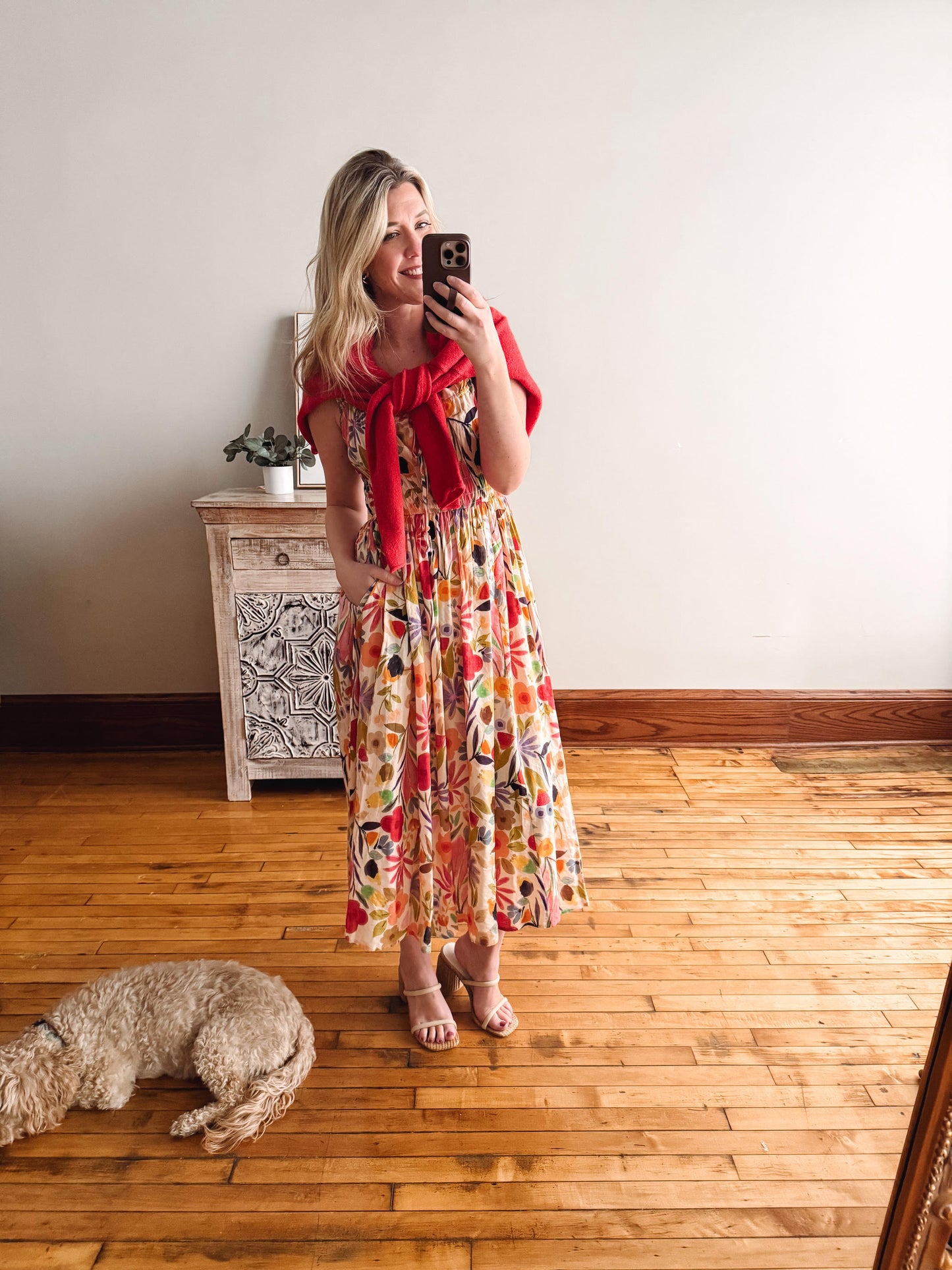 Woman in a colorful dress with a red shawl taking a mirror selfie in a room with wooden flooring and a small dog.