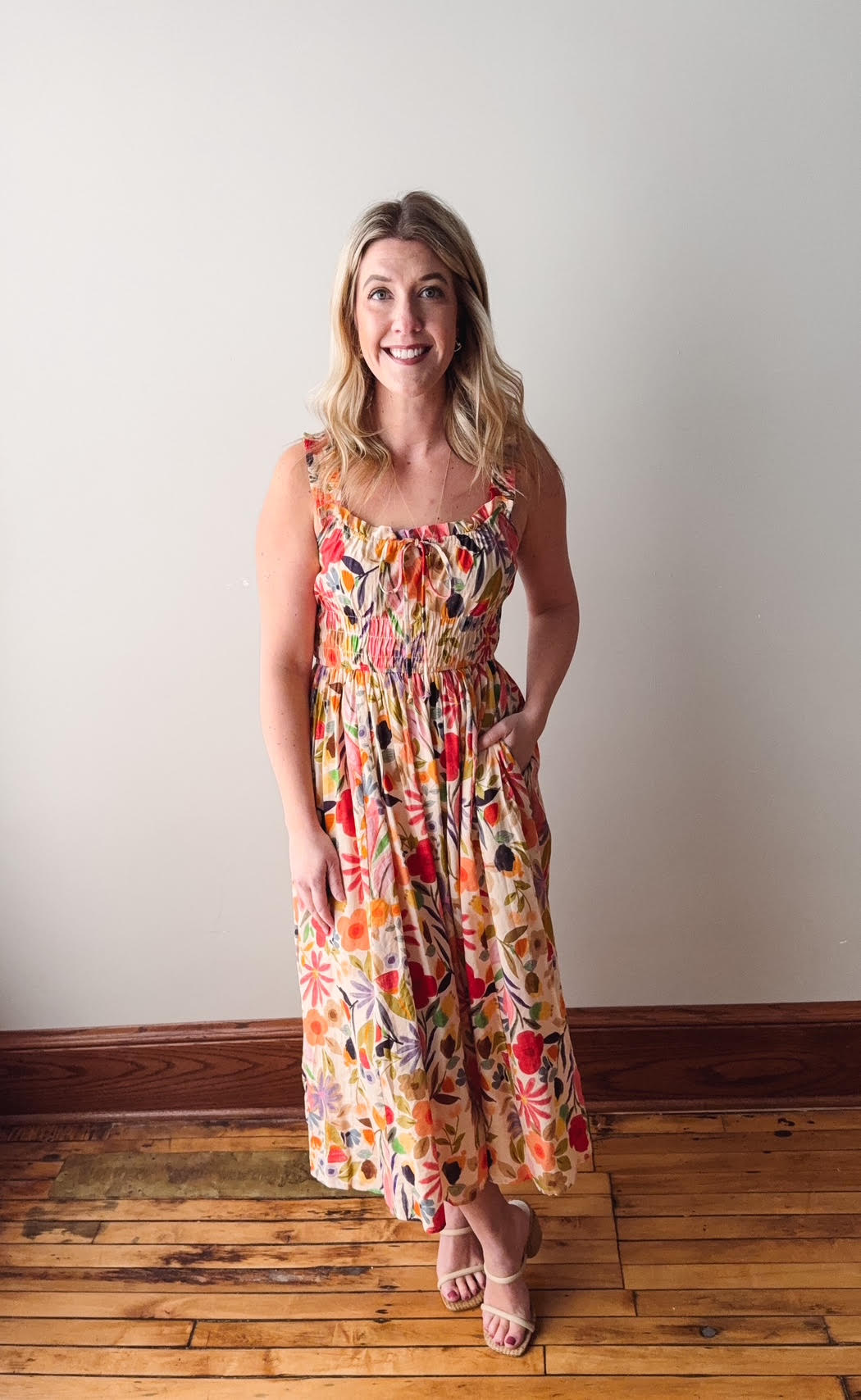 Woman wearing a colorful floral dress standing on a wooden floor against a plain wall.
