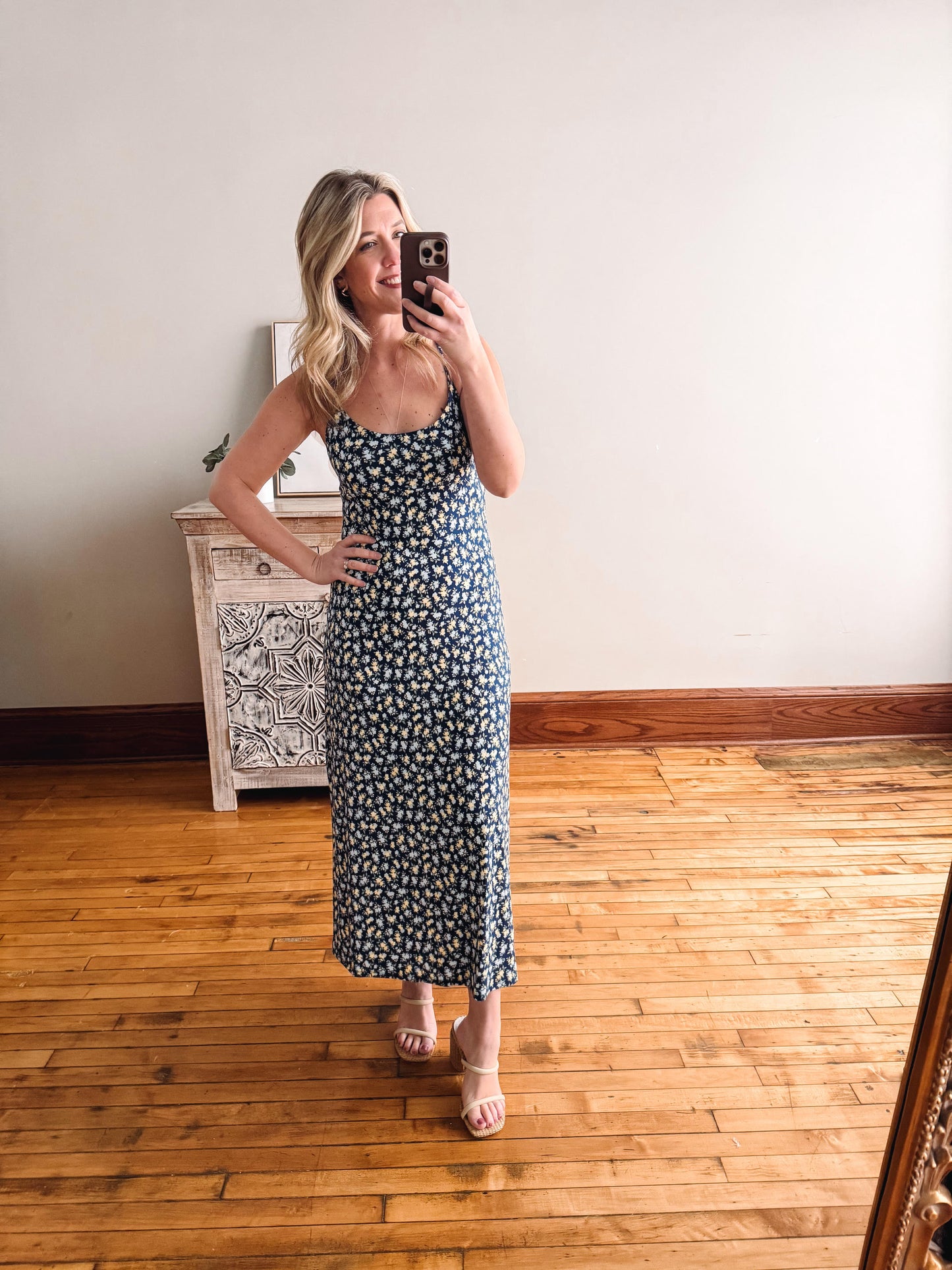 Woman taking a mirror selfie wearing a floral dress in a room with wooden flooring.