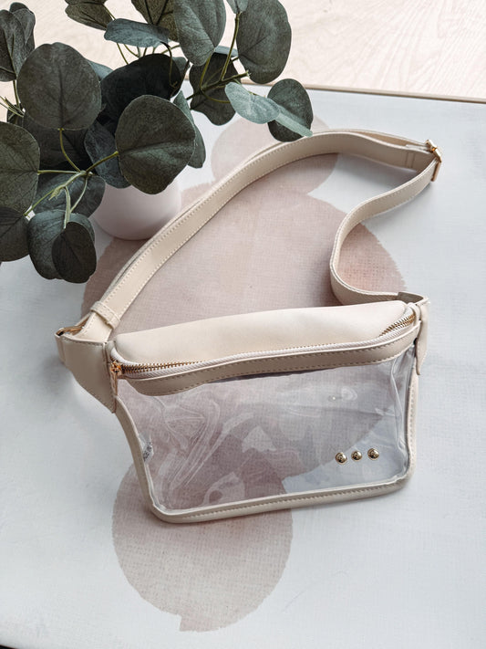 Beige crossbody bag with clear front pocket on a light surface with green leaves in the background