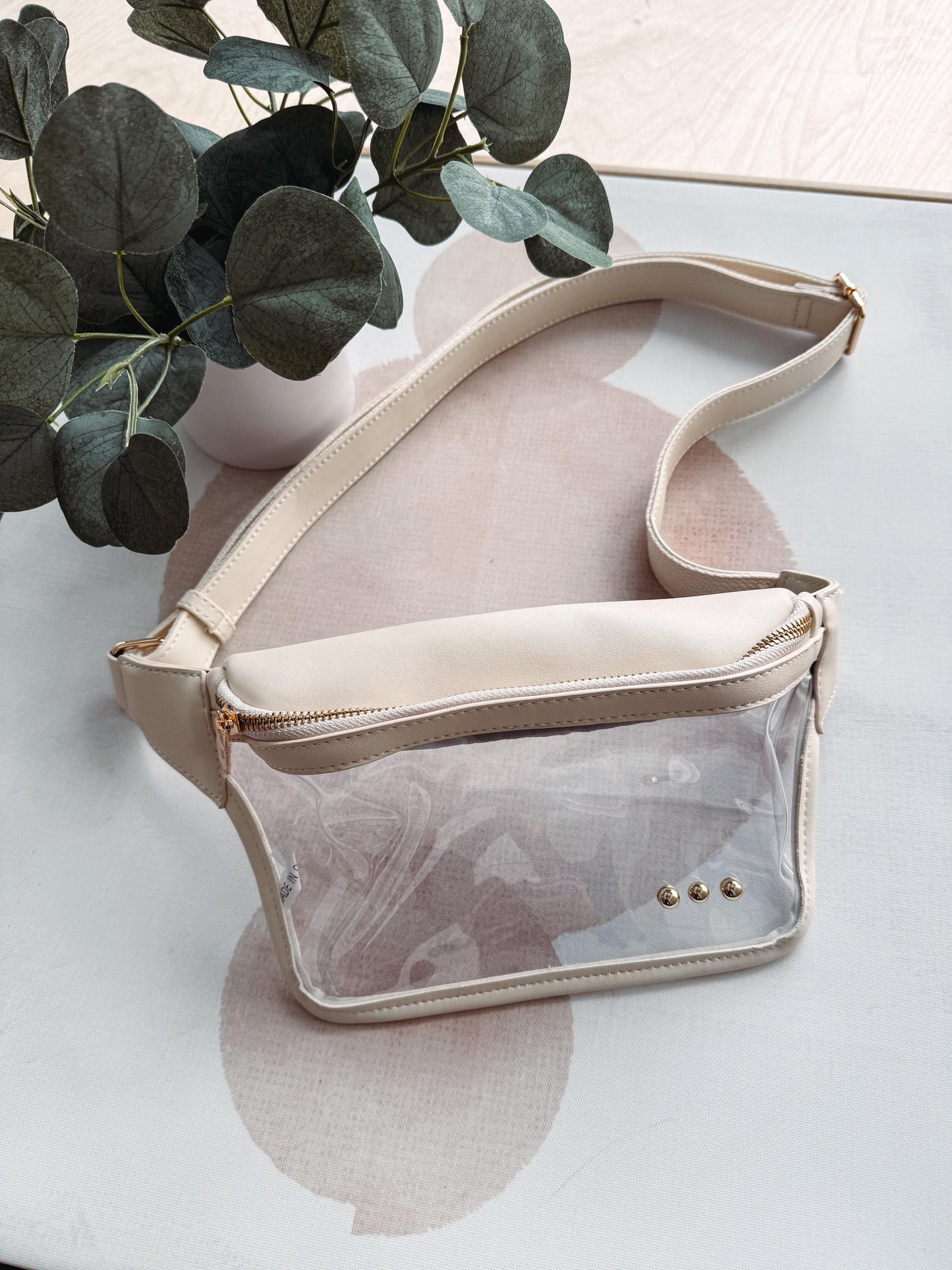 Beige crossbody bag with clear front pocket on a light surface with green leaves in the background