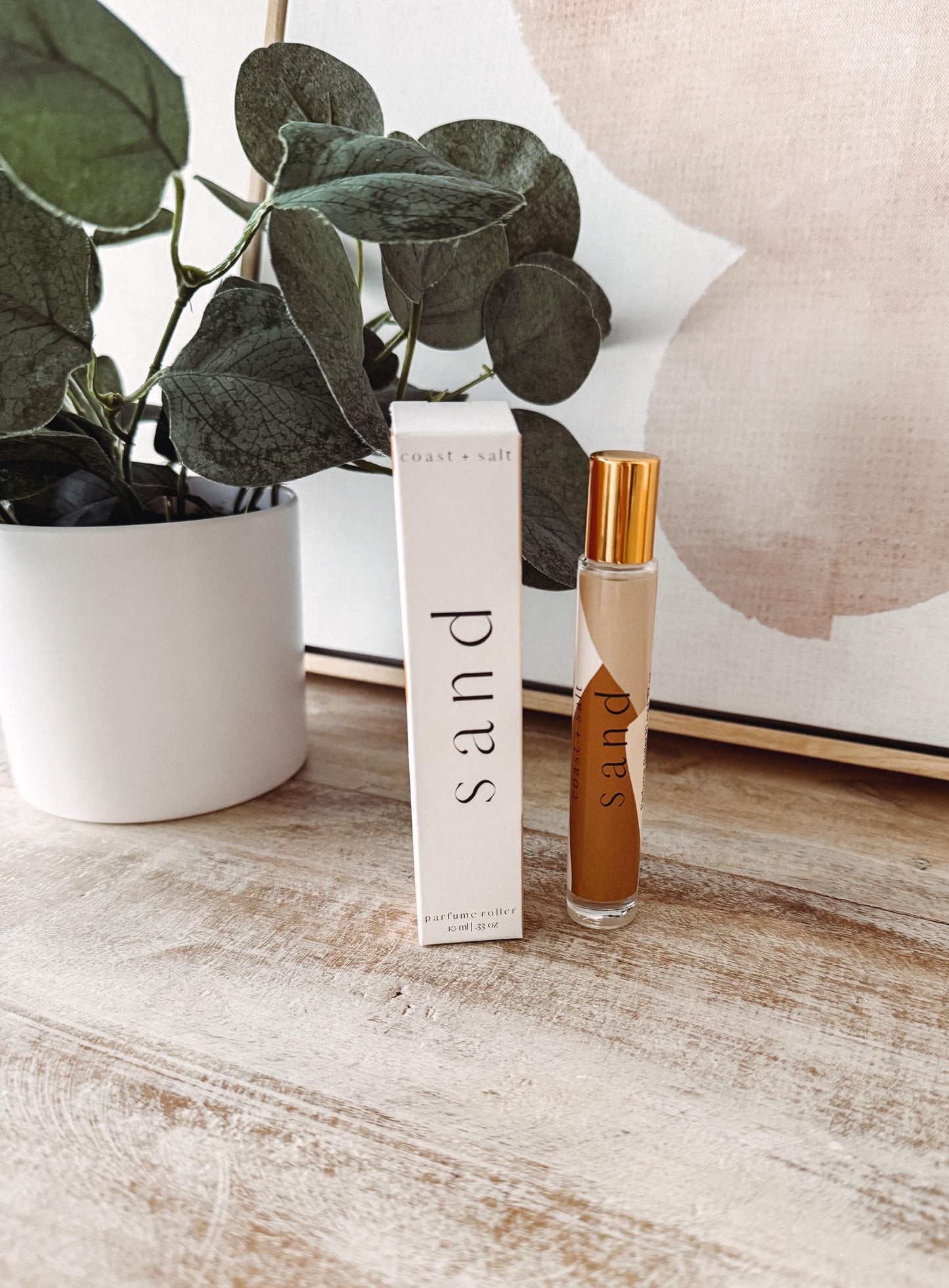Perfume bottle and packaging labeled 'sand' on a wooden surface with a plant in the background.