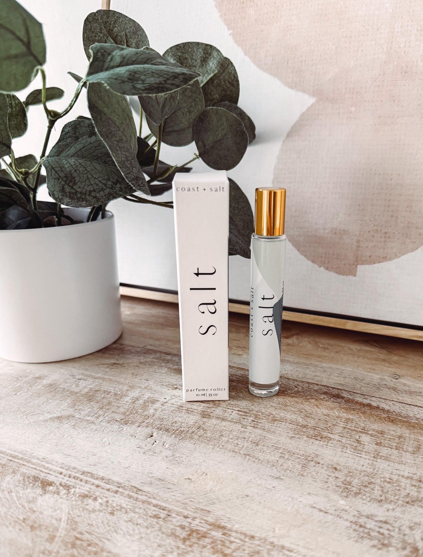 Small bottle of 'salt' perfume on a wooden surface with a plant and decorative item in the background.