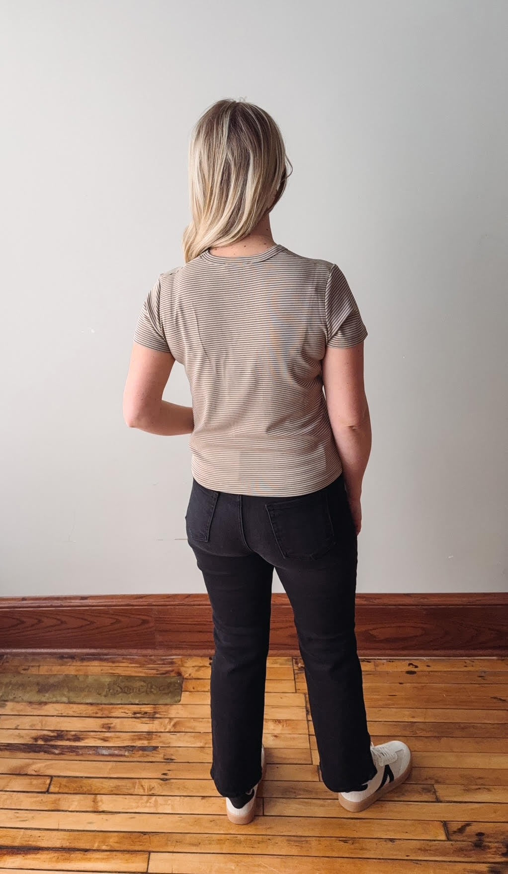 Person wearing a beige t-shirt and black pants standing on a wooden floor with a white wall background