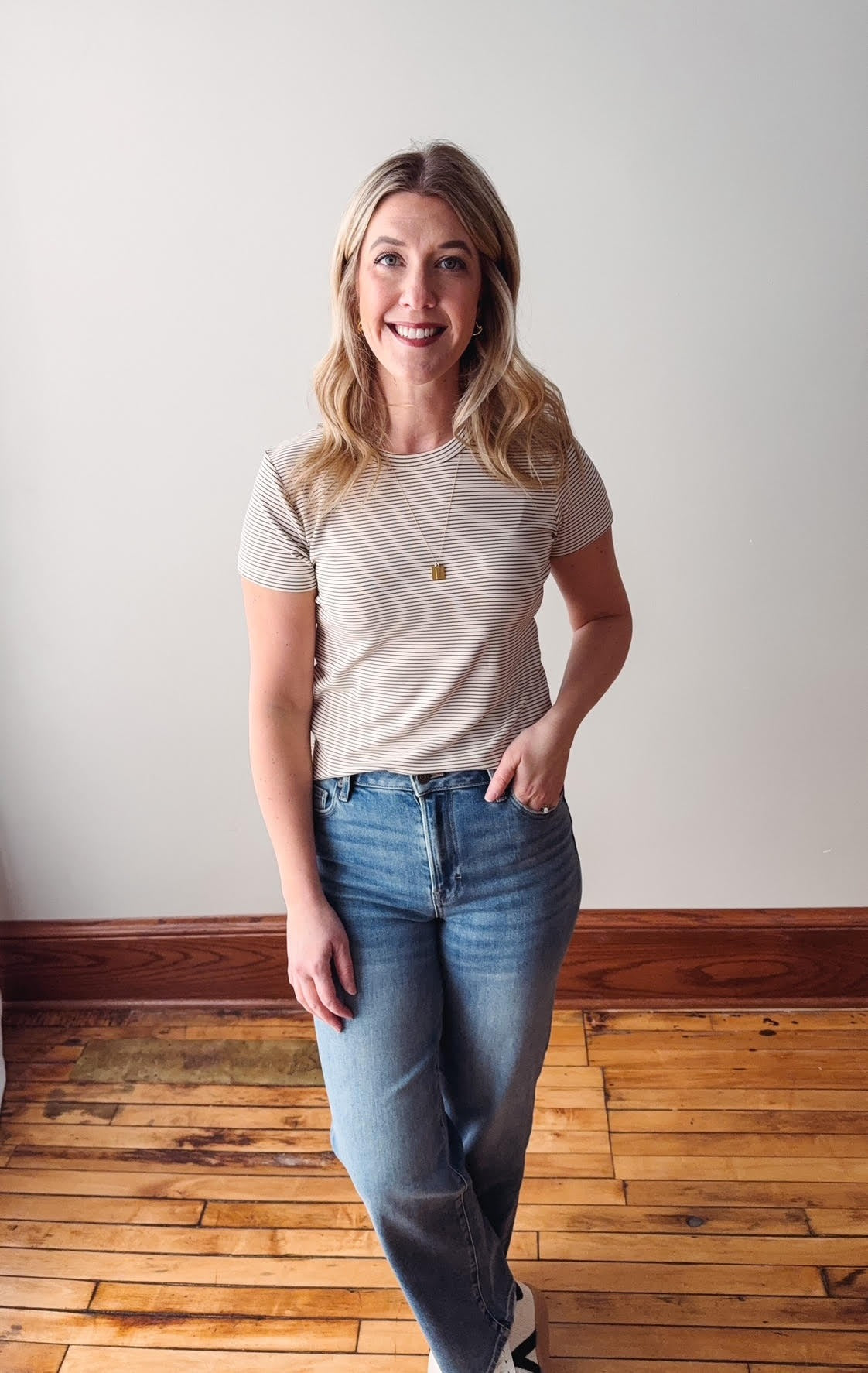 Woman wearing a striped shirt and jeans standing on a wooden floor with a white wall background