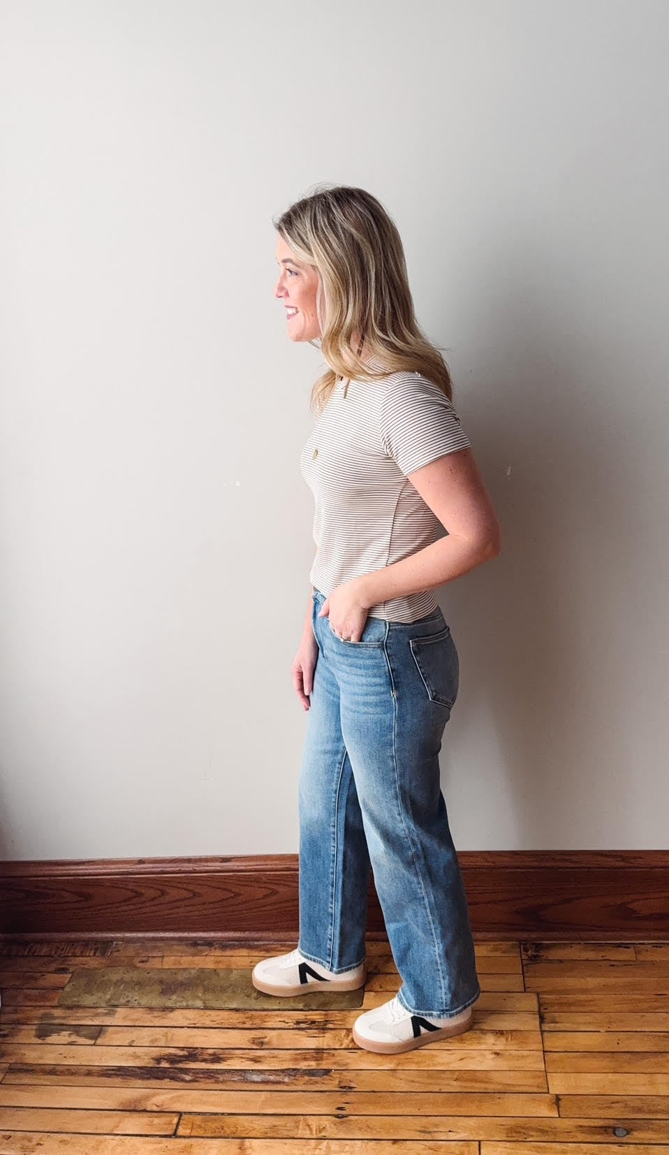 Woman standing against a white wall wearing a light gray t-shirt and blue jeans.