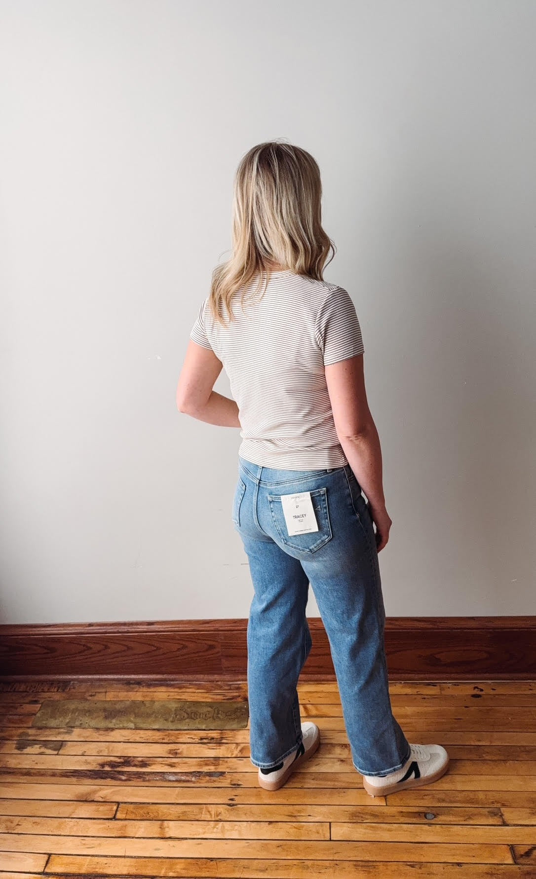 Person wearing blue jeans and a beige shirt standing on a wooden floor with a white wall background