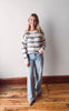 Woman wearing a striped sweater and jeans standing on a wooden floor with a white wall background