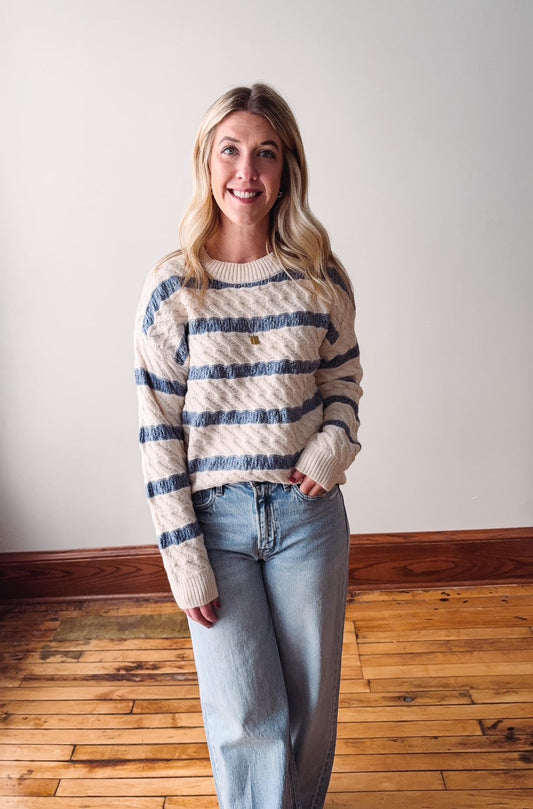 Woman wearing a striped sweater and jeans standing on a wooden floor with a white wall background