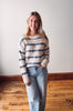 Woman wearing a striped sweater and jeans standing on a wooden floor with a white wall background