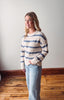 Woman wearing a striped sweater and jeans standing in a room with wooden flooring and a white wall.