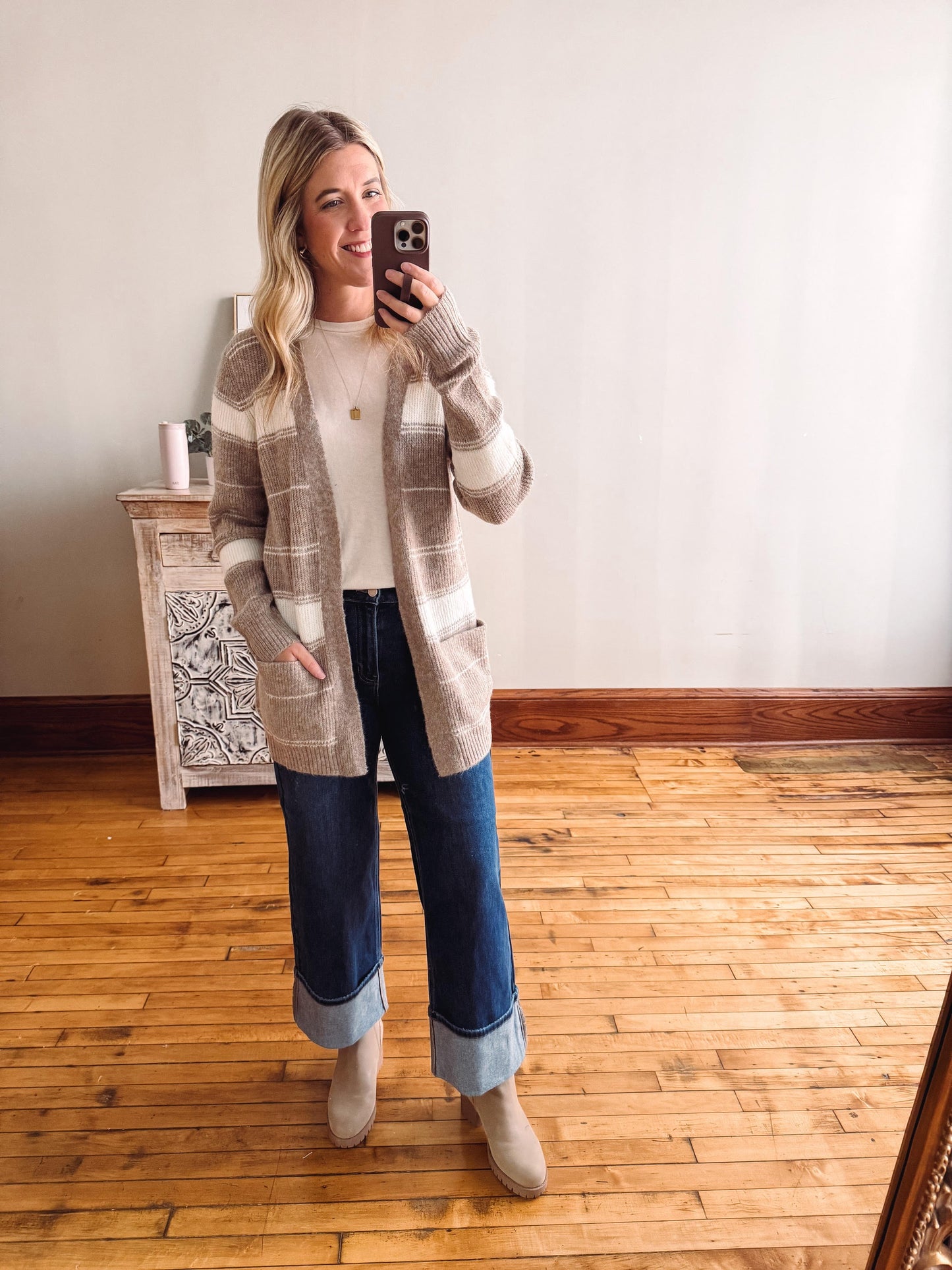 Woman taking a mirror selfie wearing a long plaid cardigan, white shirt, blue jeans, and beige boots.