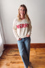 Woman wearing a white sweater with 'MERRY' in red text, standing in a room with wooden flooring and a white wall.