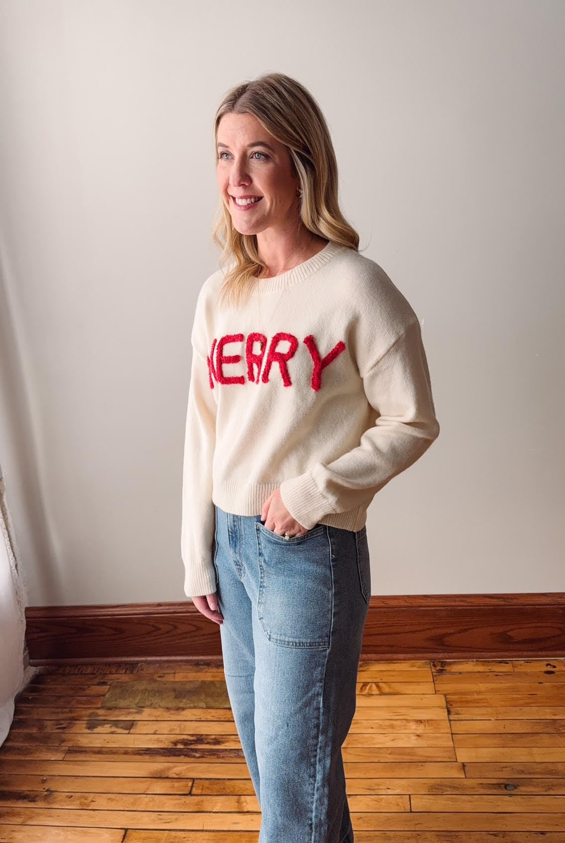 Woman wearing a cream sweater with red text standing in a room with wooden flooring and white walls.