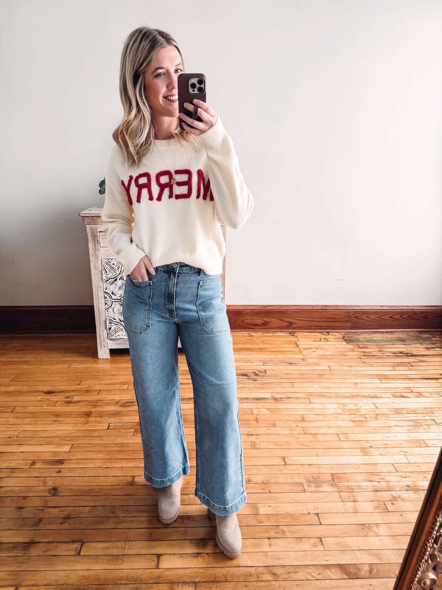 Woman taking a mirror selfie wearing a white sweater with red text and blue jeans.