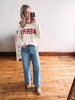 Woman taking a mirror selfie wearing a white sweater with red text and blue jeans.