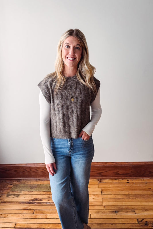 Woman wearing a sleeveless checkered top and jeans standing on a wooden floor with a white wall background.