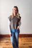 Woman wearing a sleeveless checkered top and jeans standing on a wooden floor with a white wall background.