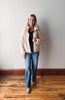 Woman wearing a beige fleece jacket and blue jeans standing against a plain wall.