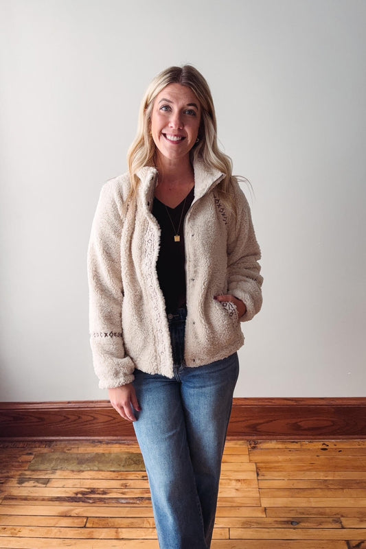 Woman wearing a beige sherpa jacket and blue jeans standing against a plain wall.