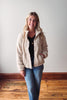 Woman wearing a beige sherpa jacket and blue jeans standing against a plain wall.