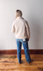 Person wearing a light pink jacket and blue jeans standing against a plain wall.
