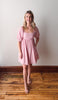 Woman wearing a pink dress standing on a wooden floor with a white wall background
