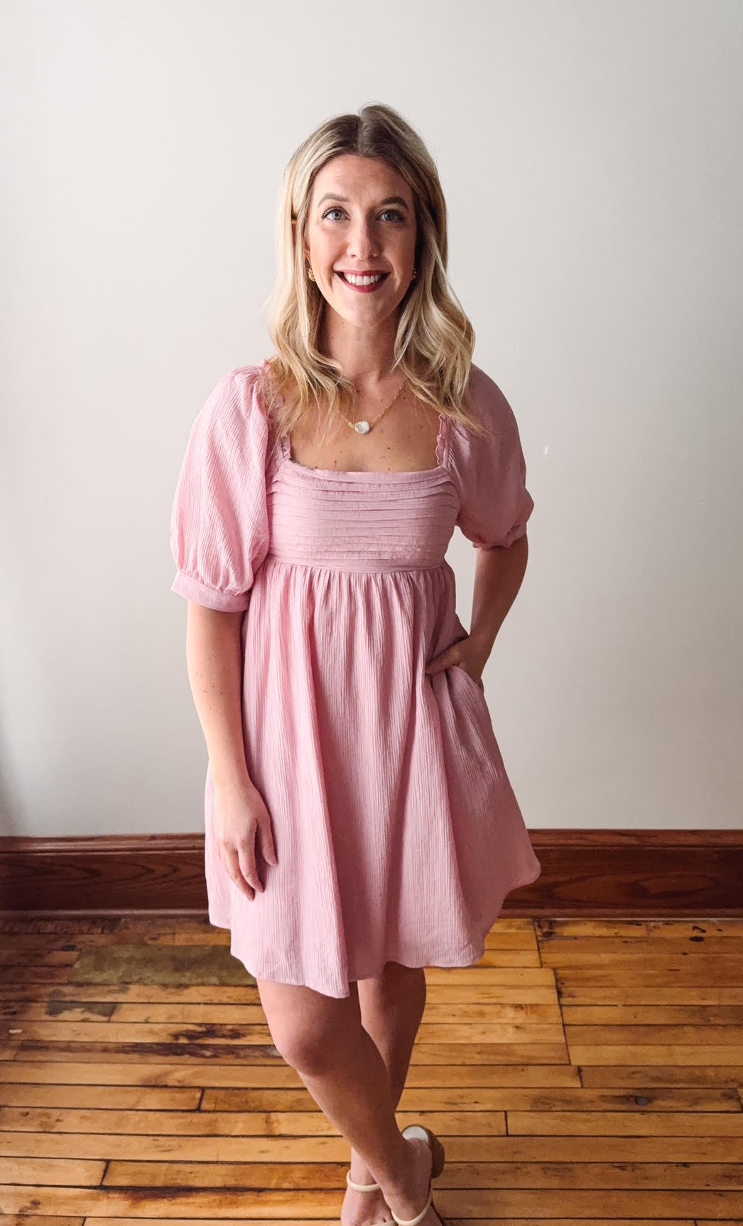Woman wearing a pink dress standing on a wooden floor with a white wall background