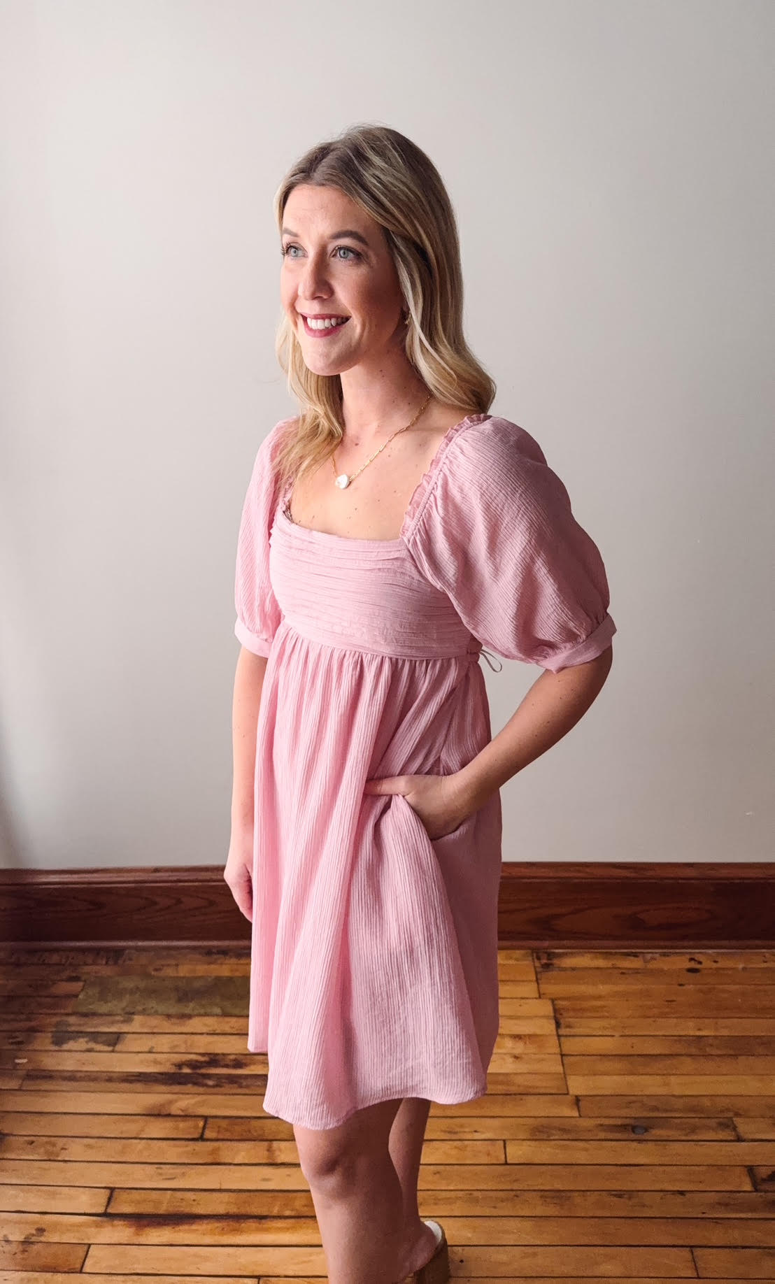 Woman wearing a pink dress standing on a wooden floor with a white wall background