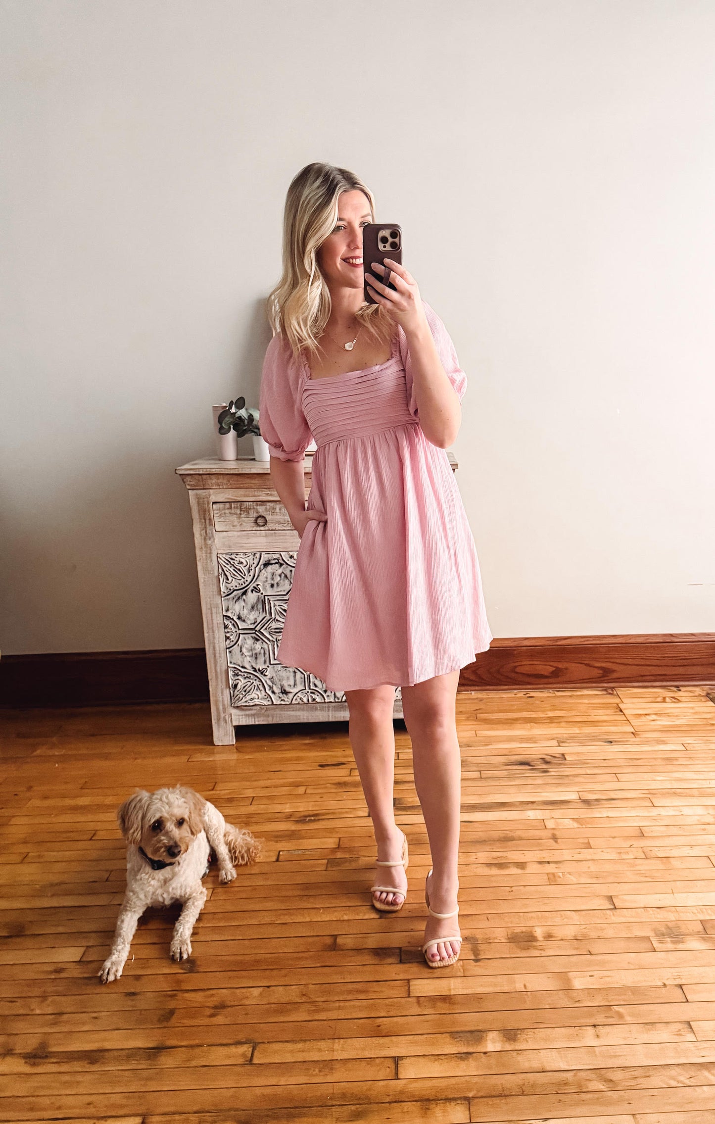 Woman in a pink dress taking a mirror selfie with a dog on a wooden floor.