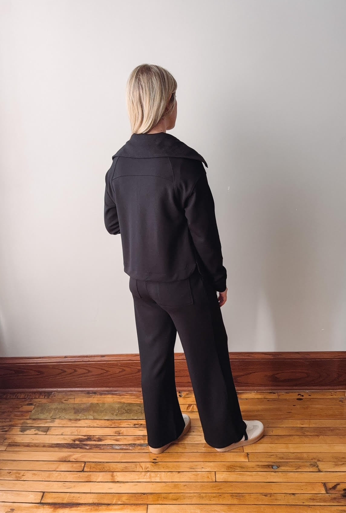 Person wearing a black outfit standing on a wooden floor with a white wall background